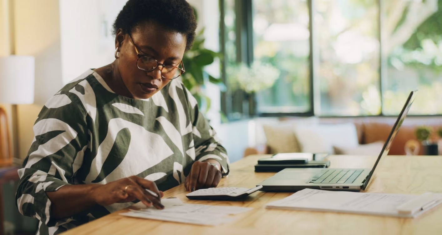Woman wearing glasses and a patterned sweater focused on writing on papers at a wooden table with a laptop and documents nearby in a bright room with large windows and green plants outside.