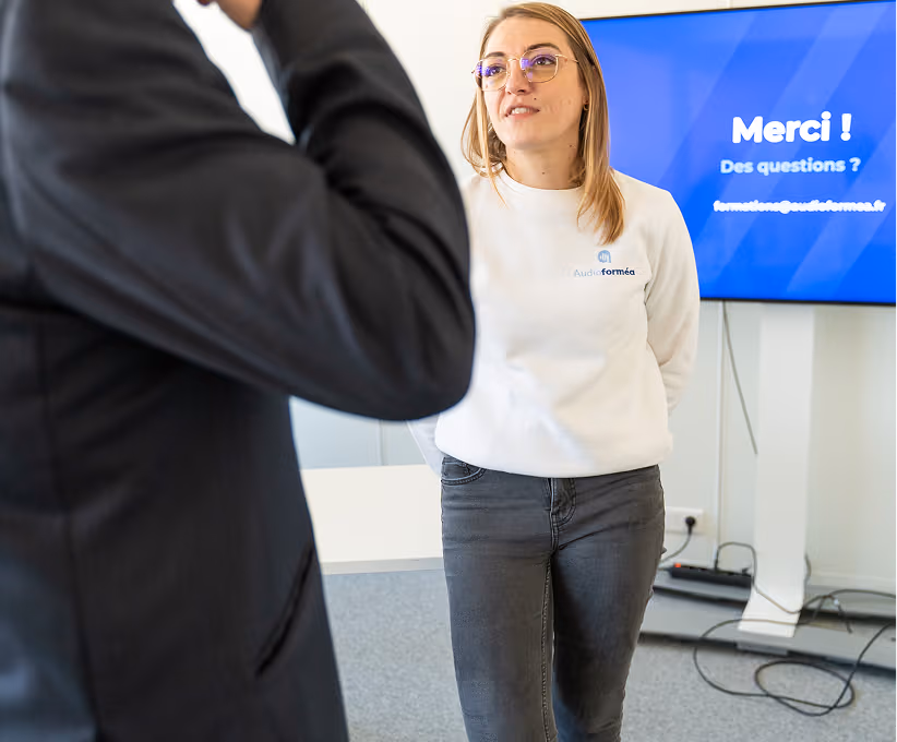 Une femme en lunettes et sweat-shirt blanc parle à un homme en costume devant un écran bleu.