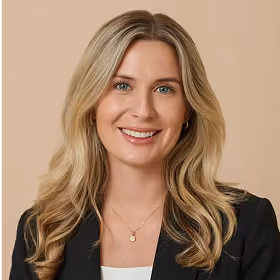 Smiling blond woman with long hair wearing a black blazer and gold necklace on beige background.