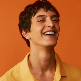 Young man smiling broadly against an orange background, wearing a yellow shirt.