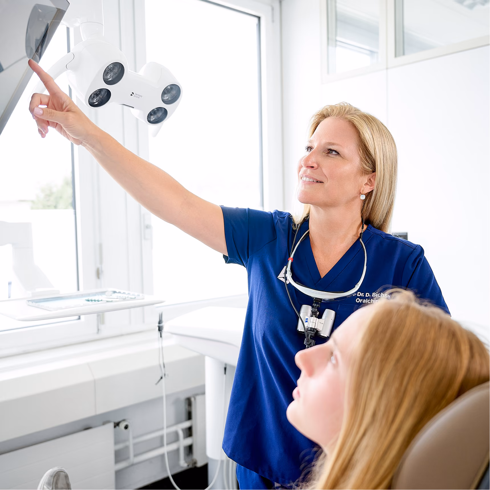 Zahnärztin zeigt auf eine Beleuchtungseinheit, während eine Patientin im Stuhl sitzt.