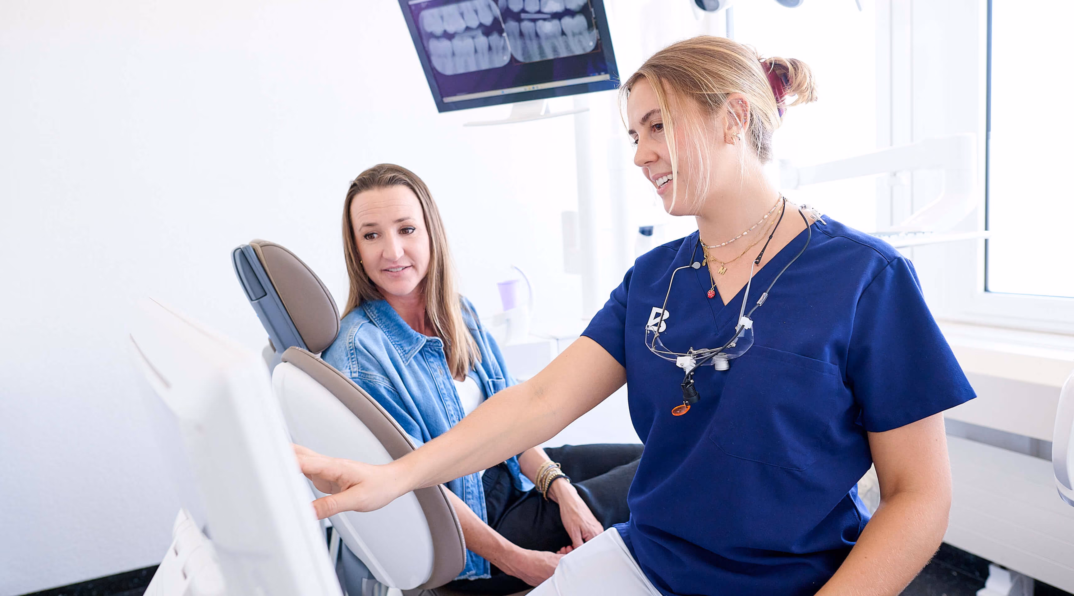 Zahnärztin zeigt Patientin im Behandlungsstuhl digitale Röntgenbilder am Computerbildschirm.
