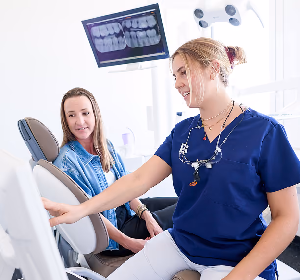 Eine Zahnärztin zeigt einer Patientin im Behandlungsstuhl am Bildschirm Röntgenaufnahmen der Zähne.