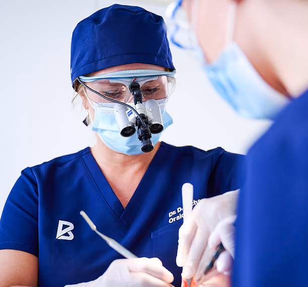 Zahnärztin mit Lupenbrille und Maske behandelt Patient im Operationssaal.