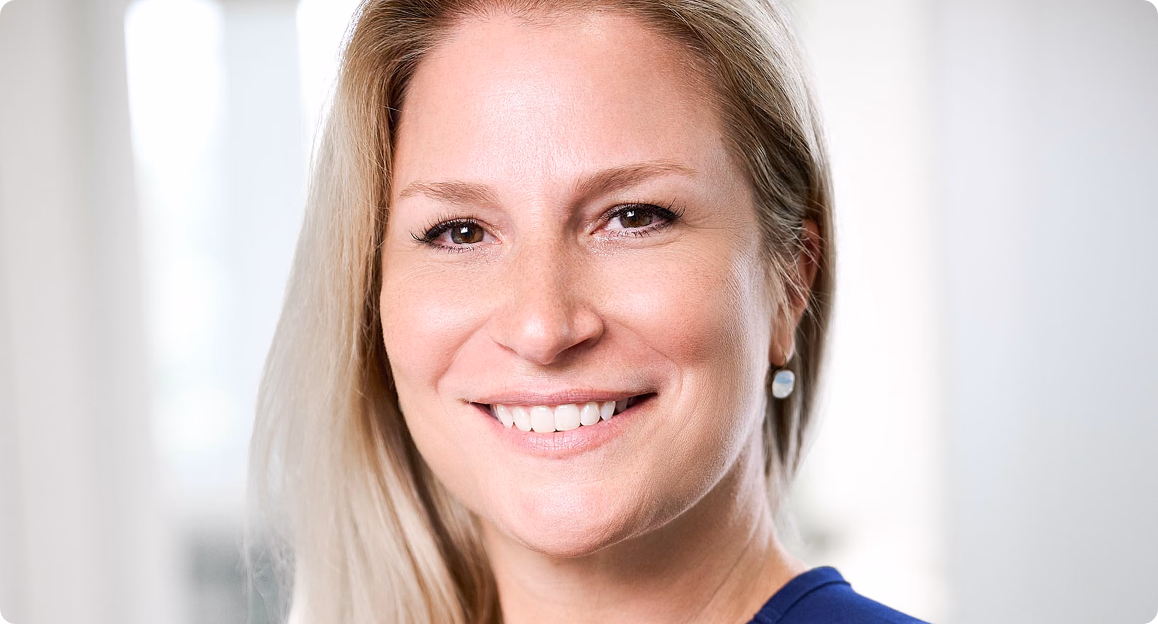 Lächelnde blonde Frau mit langen Haaren und blauem Oberteil vor hellem Hintergrund.
