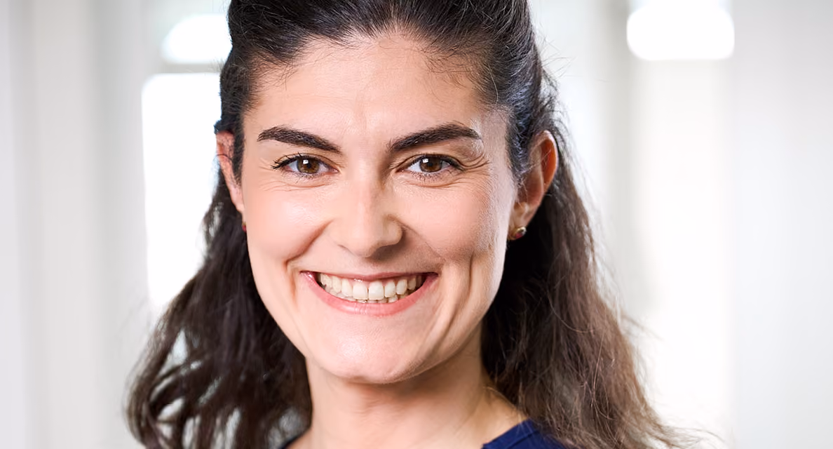 Nahaufnahme einer lächelnden Frau mit dunklem Haar und braunen Augen vor hellem Hintergrund.