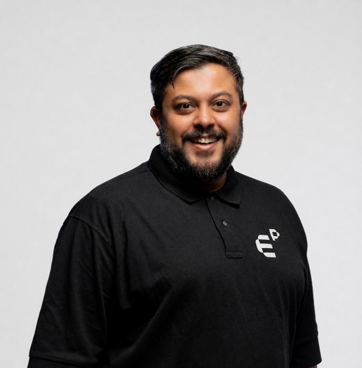 Smiling man with beard wearing a black polo shirt with a white logo on a plain background.