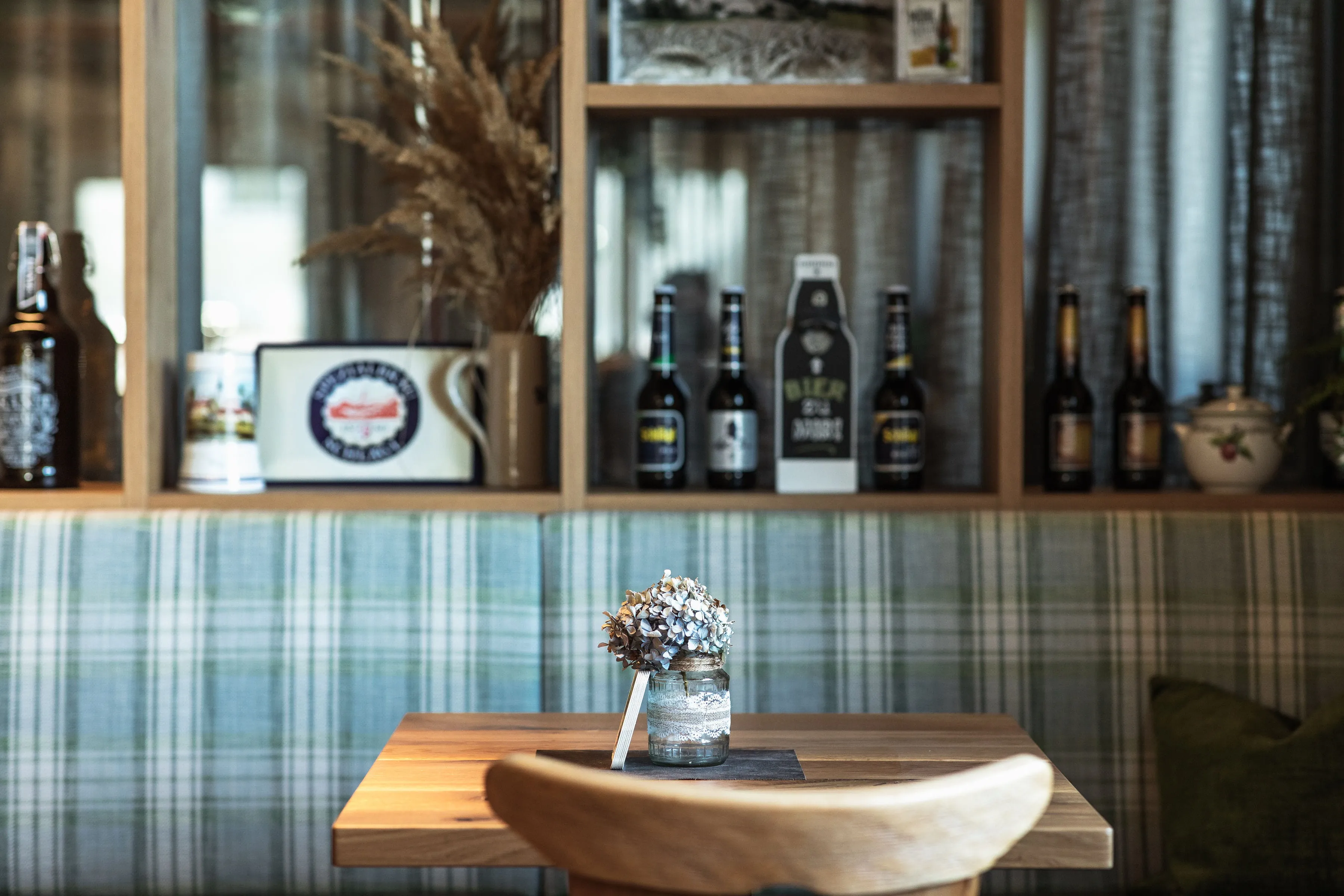 Holztisch mit getrockneten Blumen in einem Glas im Vordergrund, dahinter Regal mit Bierflaschen und Tassen.