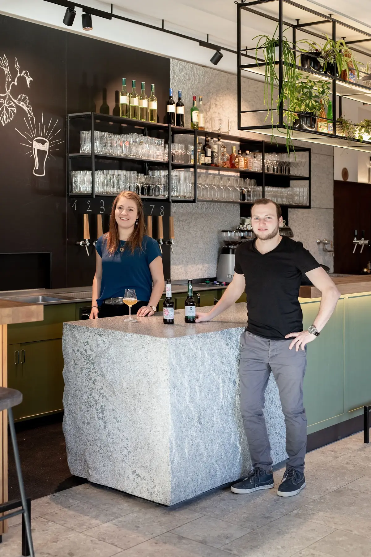 Zwei Personen stehen an einer Bar mit einem großen Steinblock als Tresen, Glas und Bierflaschen darauf.