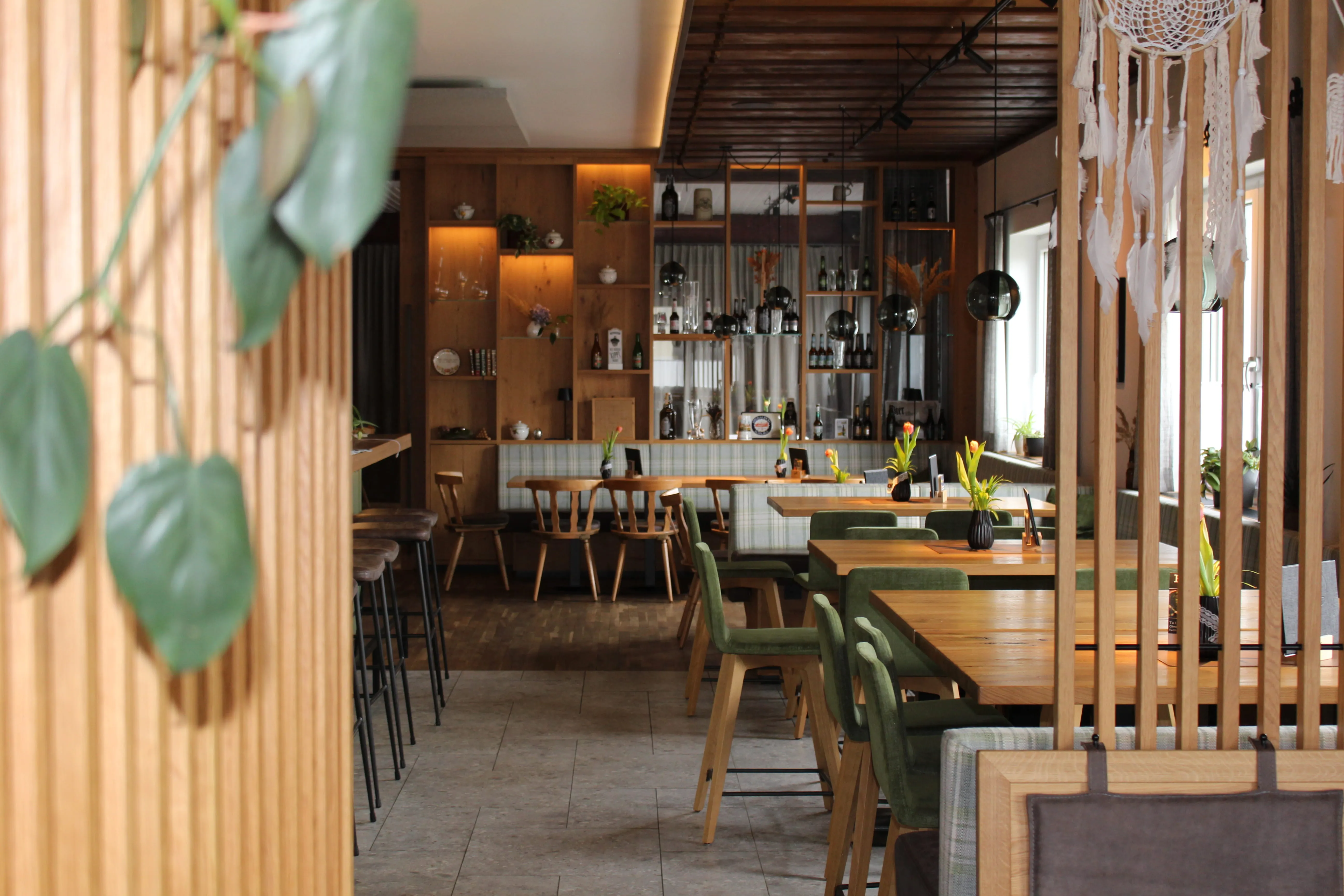 Hell eingerichtetes Restaurant mit Holzmöbeln, grünen Stühlen und dekorativen Pflanzen.
