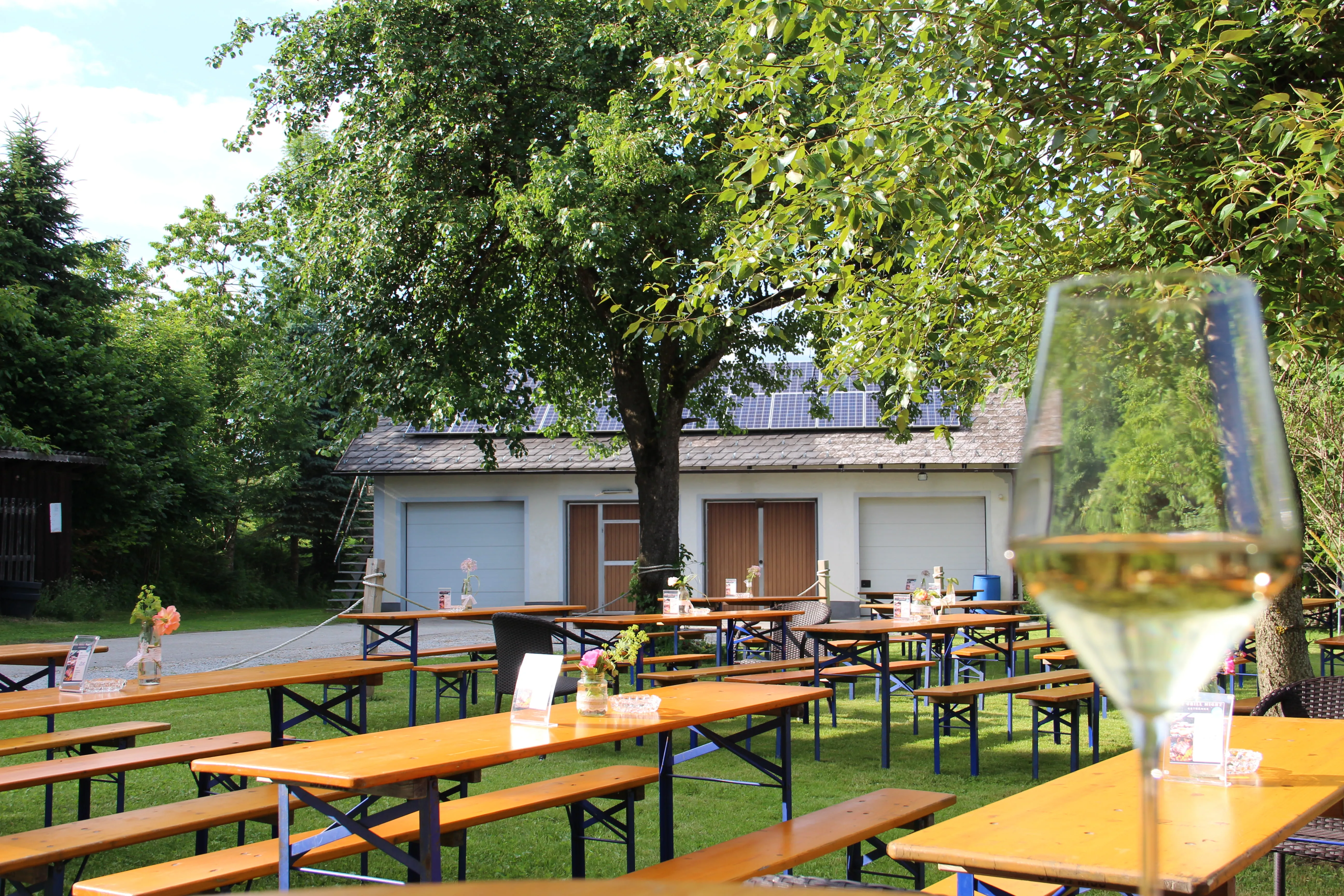 Biergartentische mit Blumenvasen und Weinglas im Vordergrund unter einem Baum vor einem Gebäude.