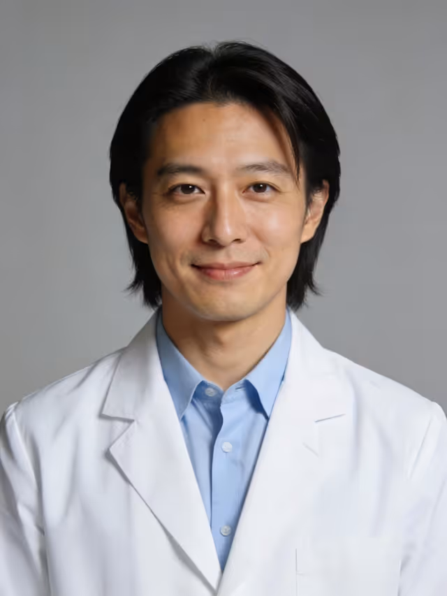 Smiling young man with black hair wearing a white coat and light blue shirt, gray background.