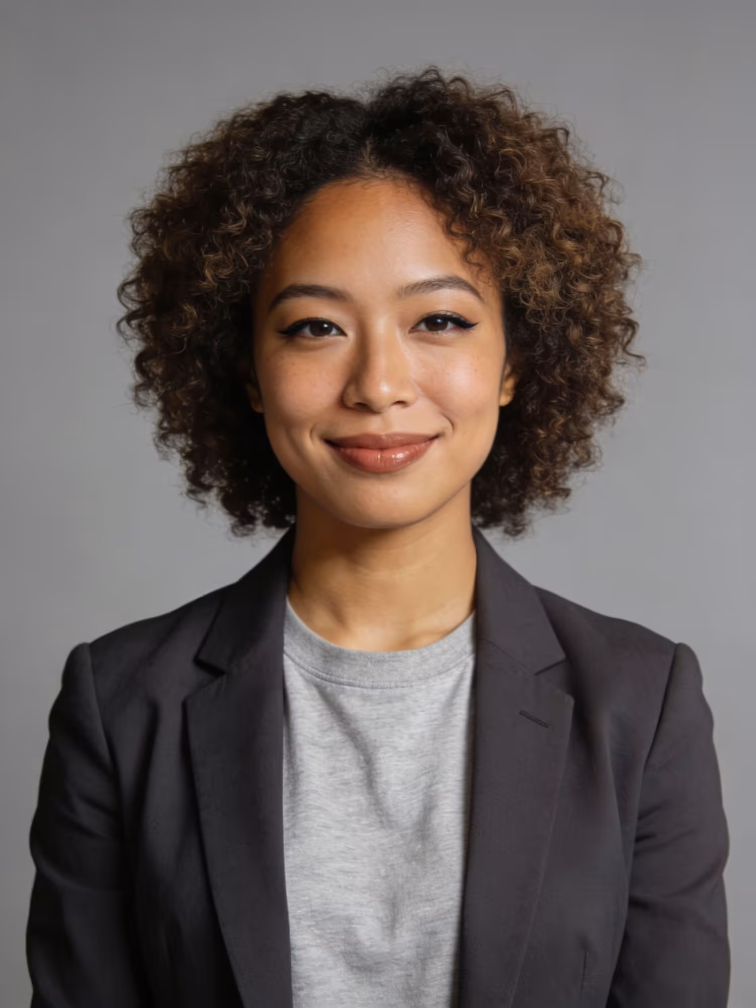 Smiling woman with curly hair wearing a gray shirt and dark blazer against gray background.