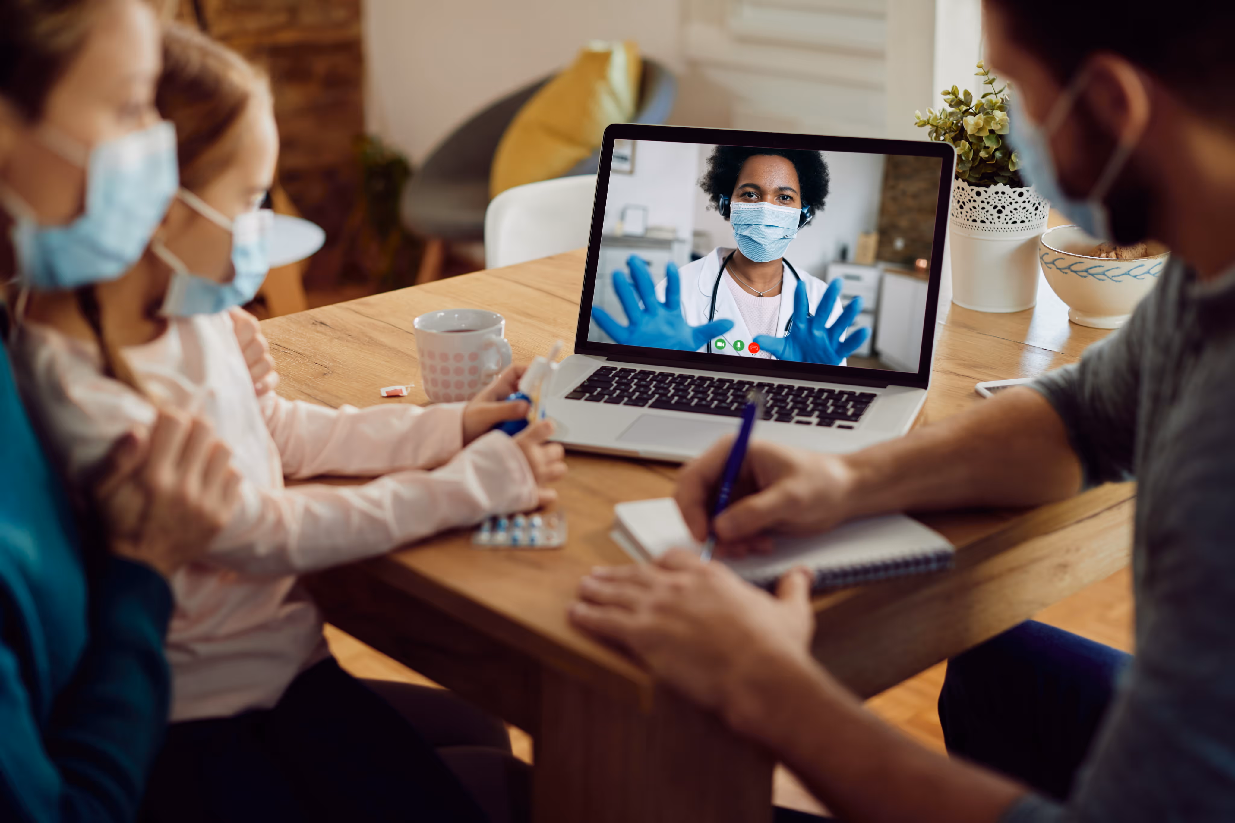 Family wears masks while consulting a masked doctor via video call on a laptop at home.