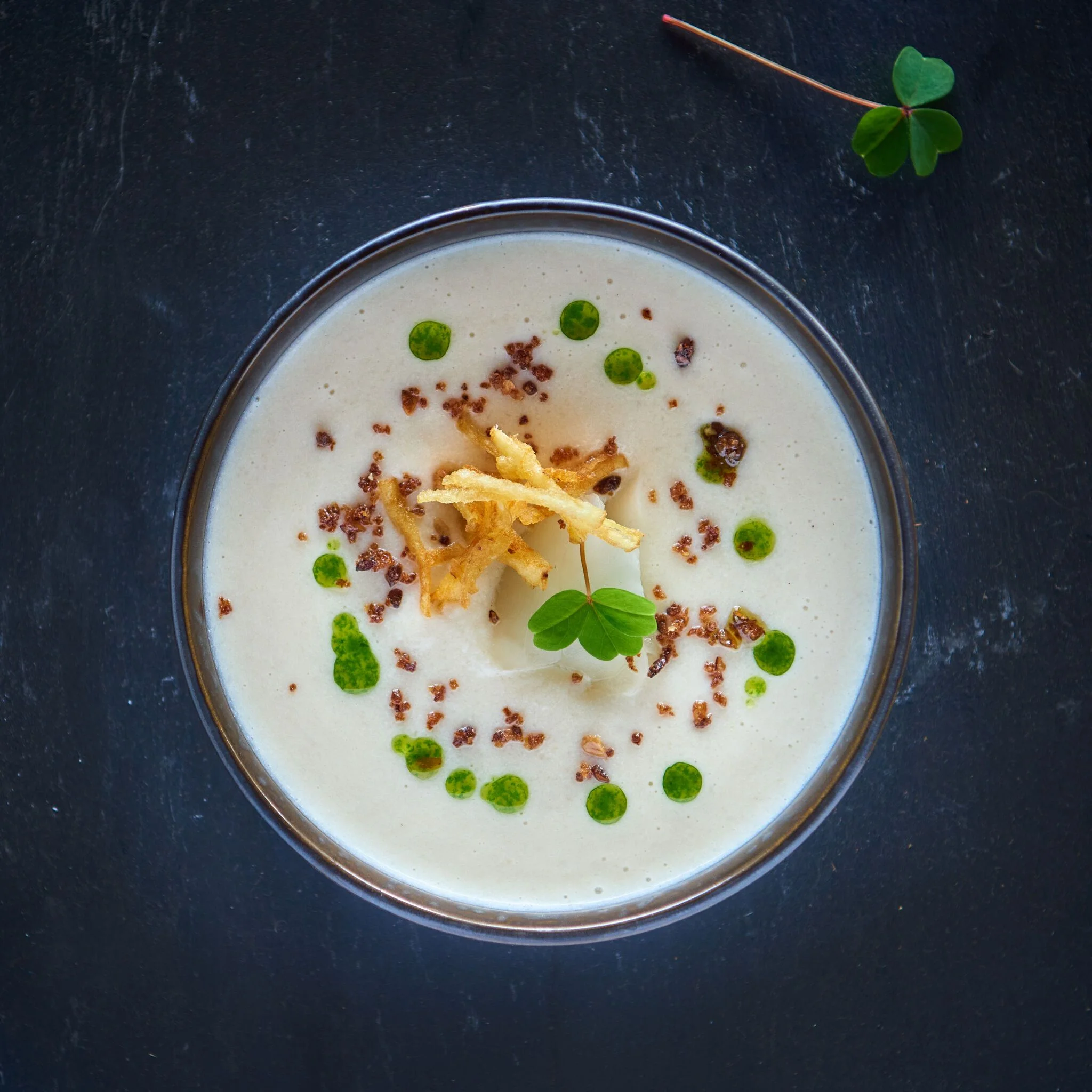 Hand pouring sauce from a small pitcher over a gourmet dish in a beige bowl on a white tablecloth.