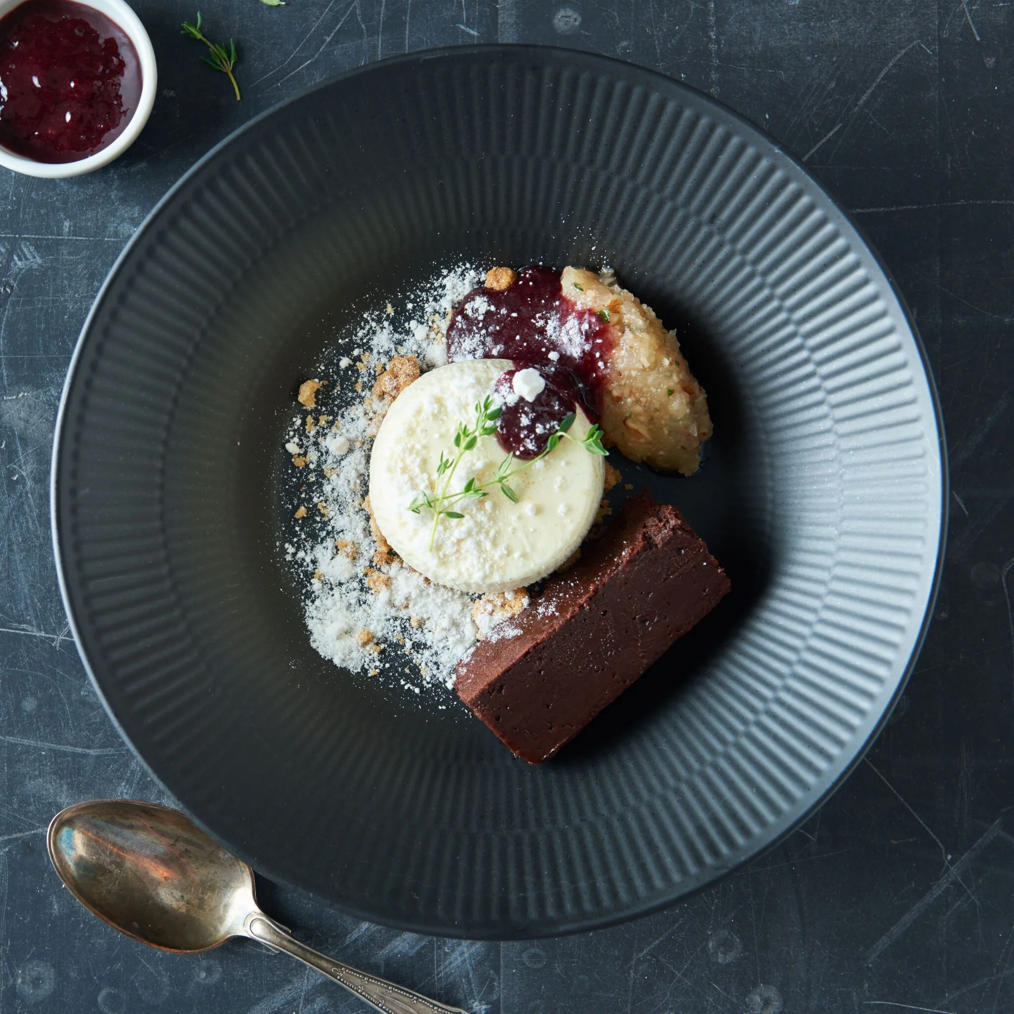 Sammensat dessert med hvid krema rørt jordbærsyltetøj og en firkantet chokoladekage på en sort tallerken.