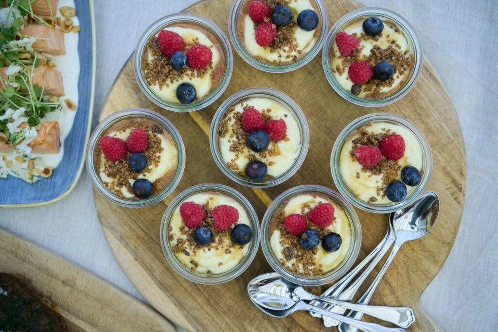 Ni glas med dessert toppet med hindbær, blåbær og knas på et træfad med skeer.
