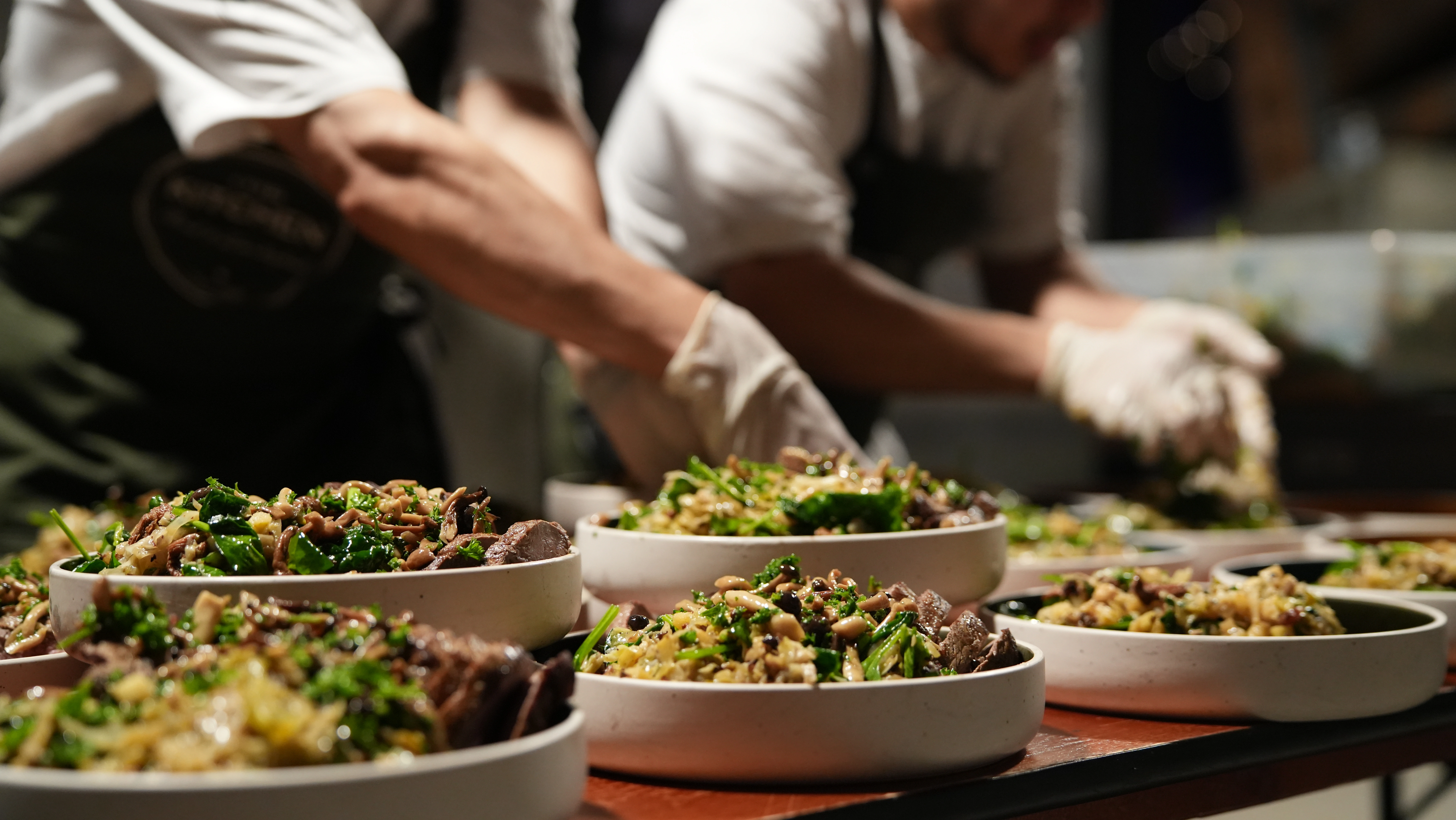 Hand pouring broth from small pitcher over a creamy pasta dish in a beige bowl on a white tablecloth.