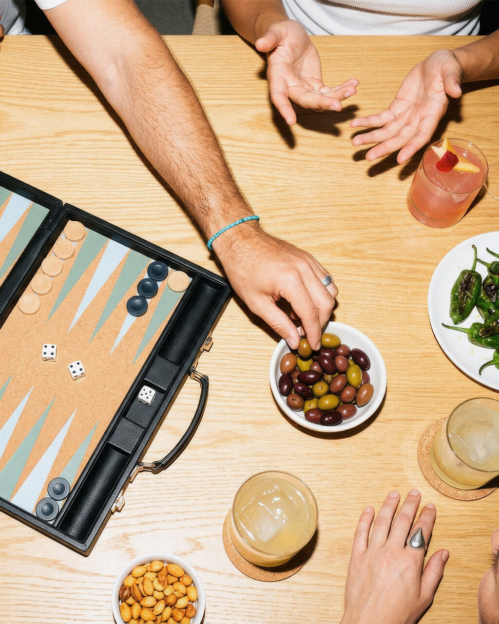 Menschen spielen Backgammon mit Snacks, Oliven, Paprika und Getränken auf einem Holztisch.