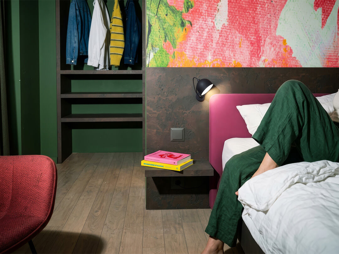 Person in grüner Hose sitzt auf Bett in modernem Zimmer mit Garderobe, rotem Sessel und Wandleuchte.