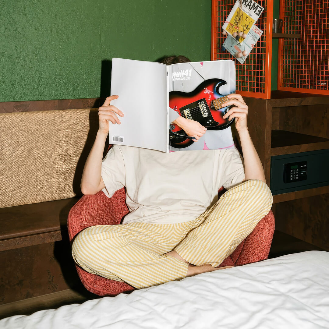Person sitzt in einem roten Sessel, liest eine Zeitschrift mit E-Gitarrenbild, Beine gekreuzt.