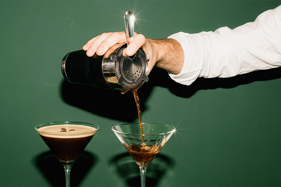 Eine Hand gießt mit einem Edelstahl-Shaker Kaffee in ein Cocktailglas vor grünem Hintergrund.