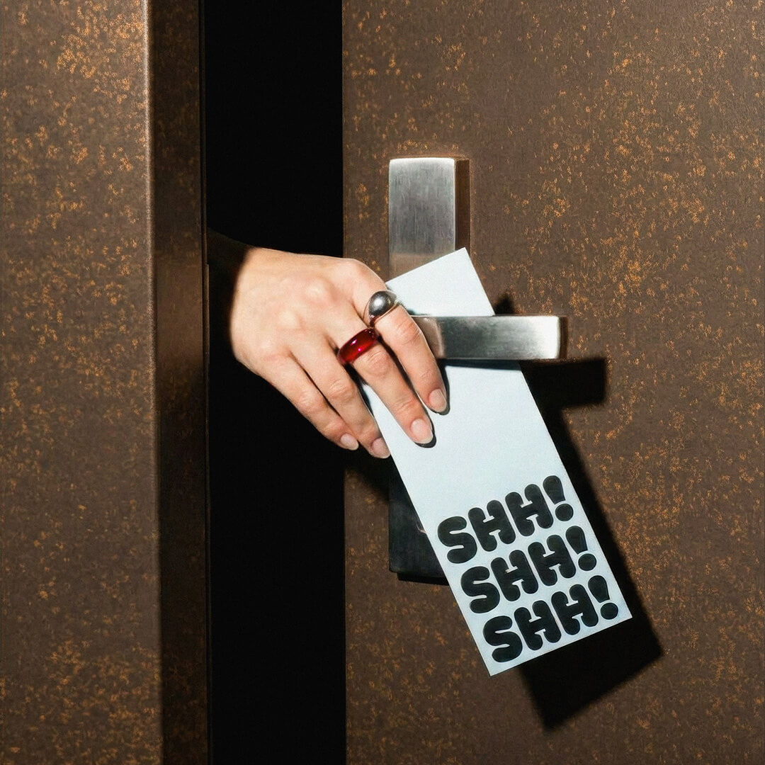 Hand with rings placing a door hanger with 'SHH! SHH! SHH!' on a brown door handle.