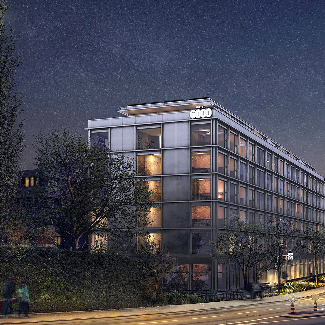 Das Hotel-6000 Gebäude bei Nacht mit beleuchteten Fenstern; auf der Strasse gehen mehrere Personen.