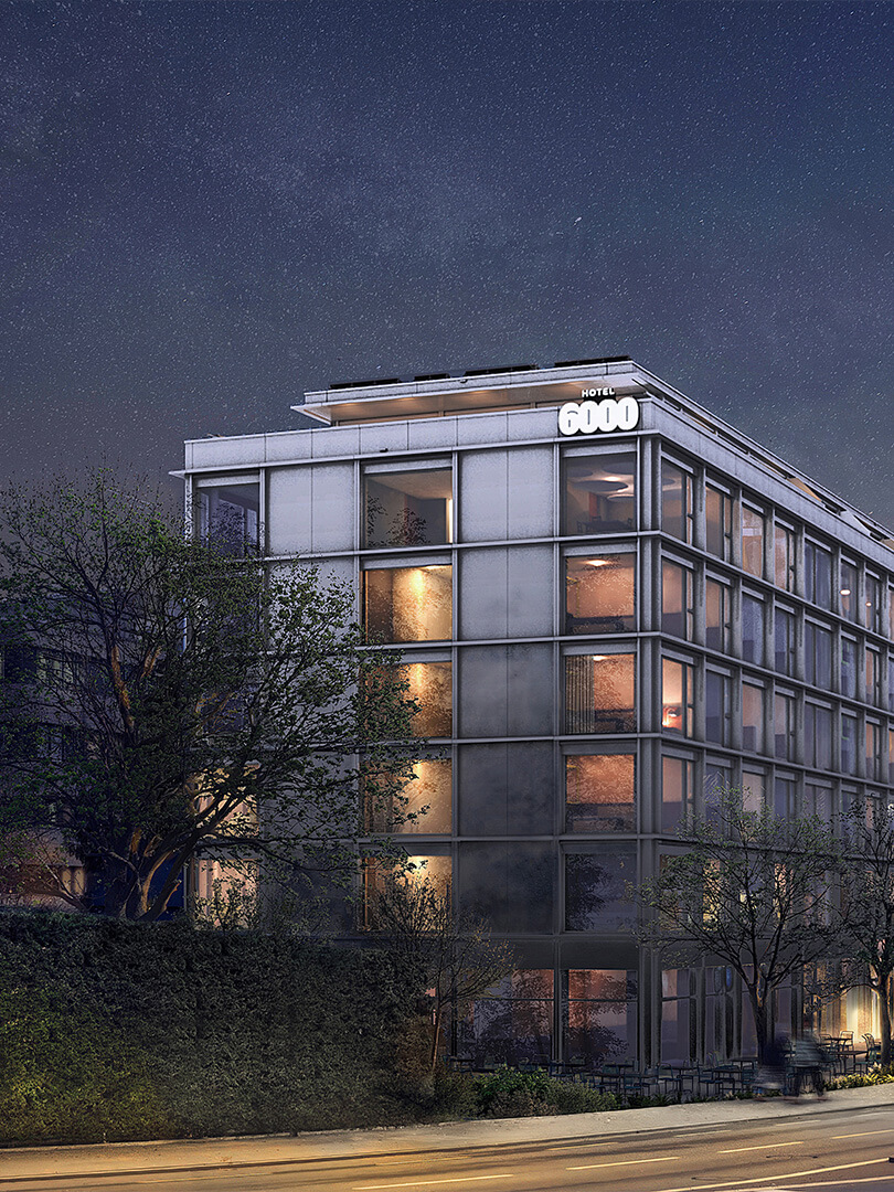 Das Hotel-6000-Gebäude bei Nacht mit beleuchteten Fenstern; auf der Strasse gehen mehrere Personen.