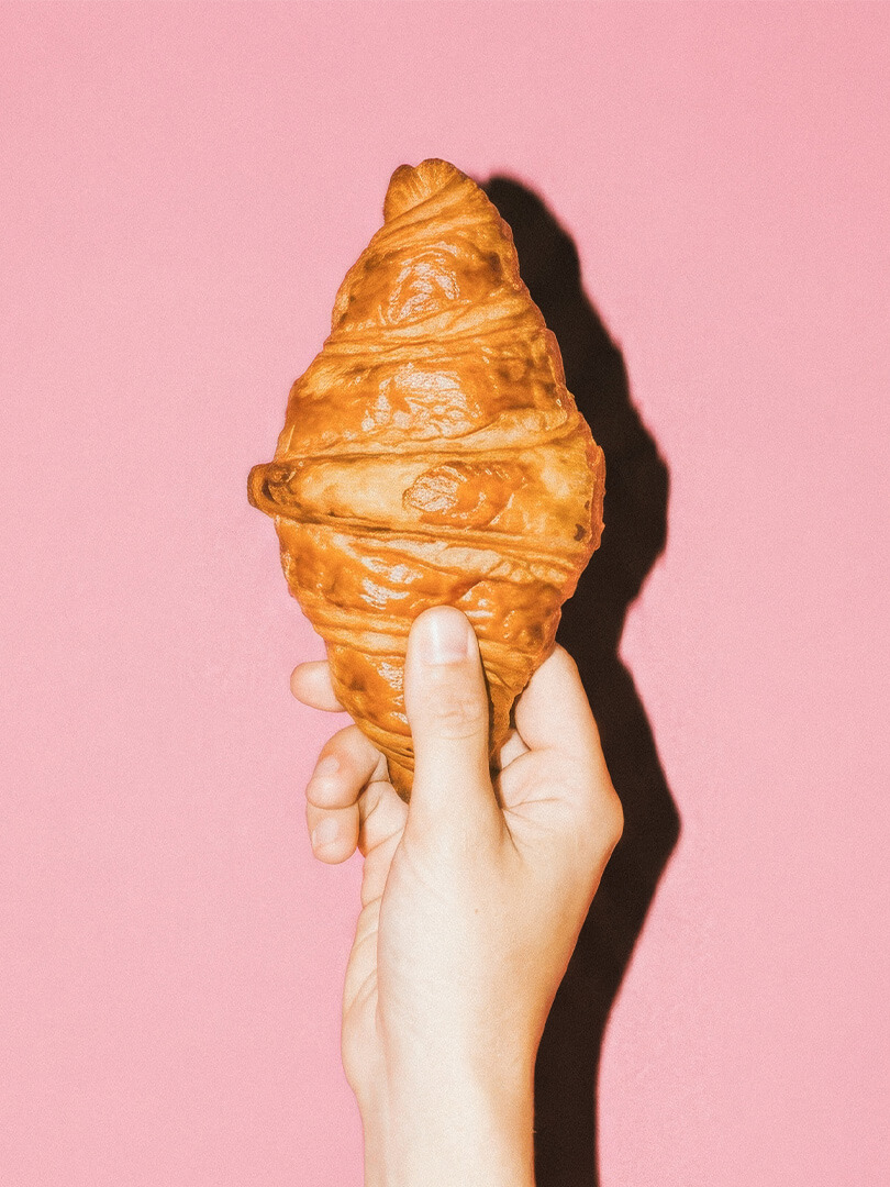 Hand hält frisch gebackenes Croissant vor rosa Hintergrund.