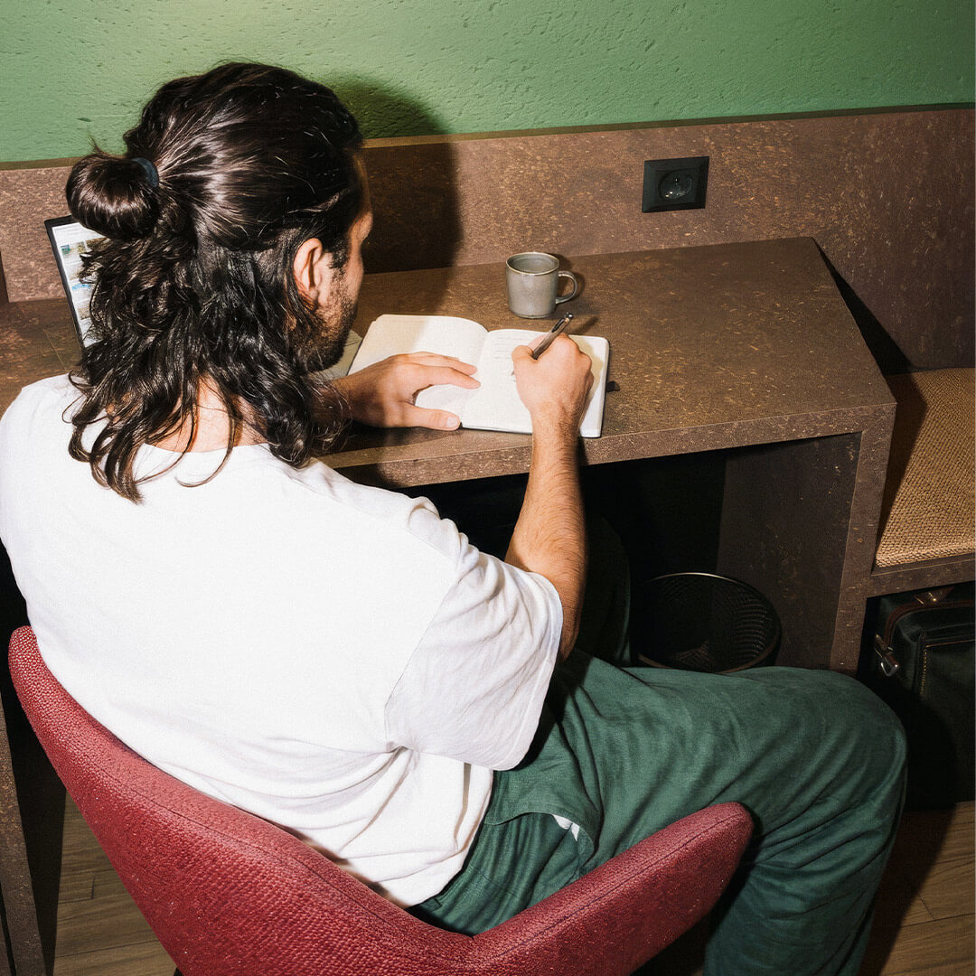 Mann mit langem Haar sitzt am Tisch in einem Zimmer im Hotel 6000 und schreibt in ein Notizbuch, daneben steht eine Kaffeetasse.