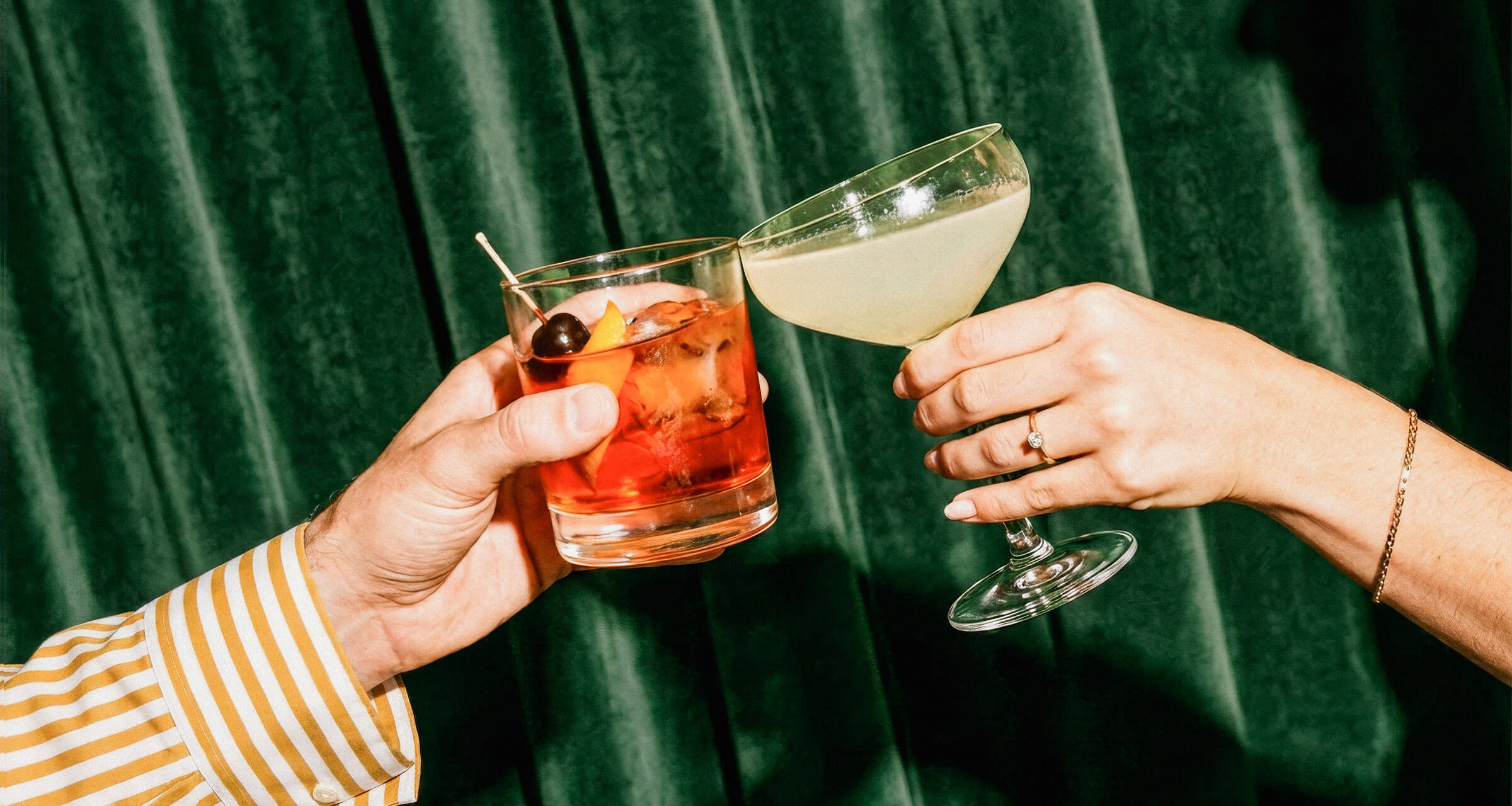 Zwei Hände stoßen mit Cocktails vor grünem Vorhang an, einer rot, einer hellgelb.