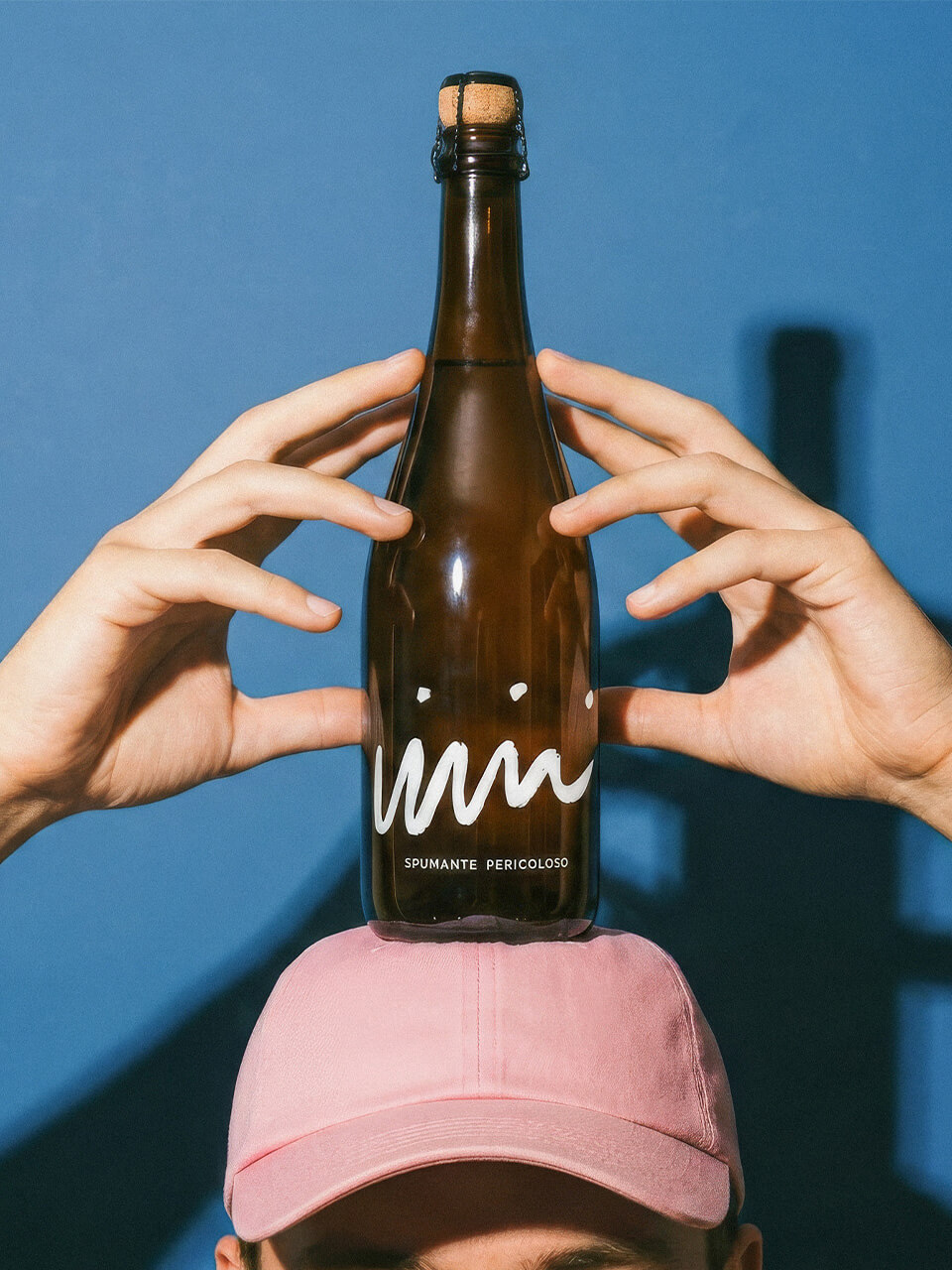 Person mit rosa Mütze balanciert eine braune Flasche mit Korken und weißer Schrift auf dem Kopf.