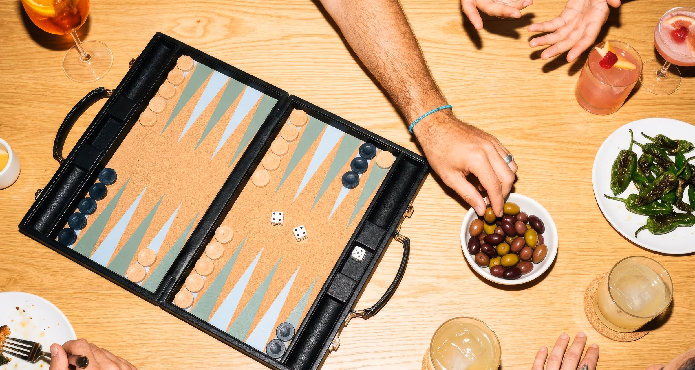 Person spielt Backgammon an einem Tisch mit Snacks, Getränken und einer Schale Oliven.