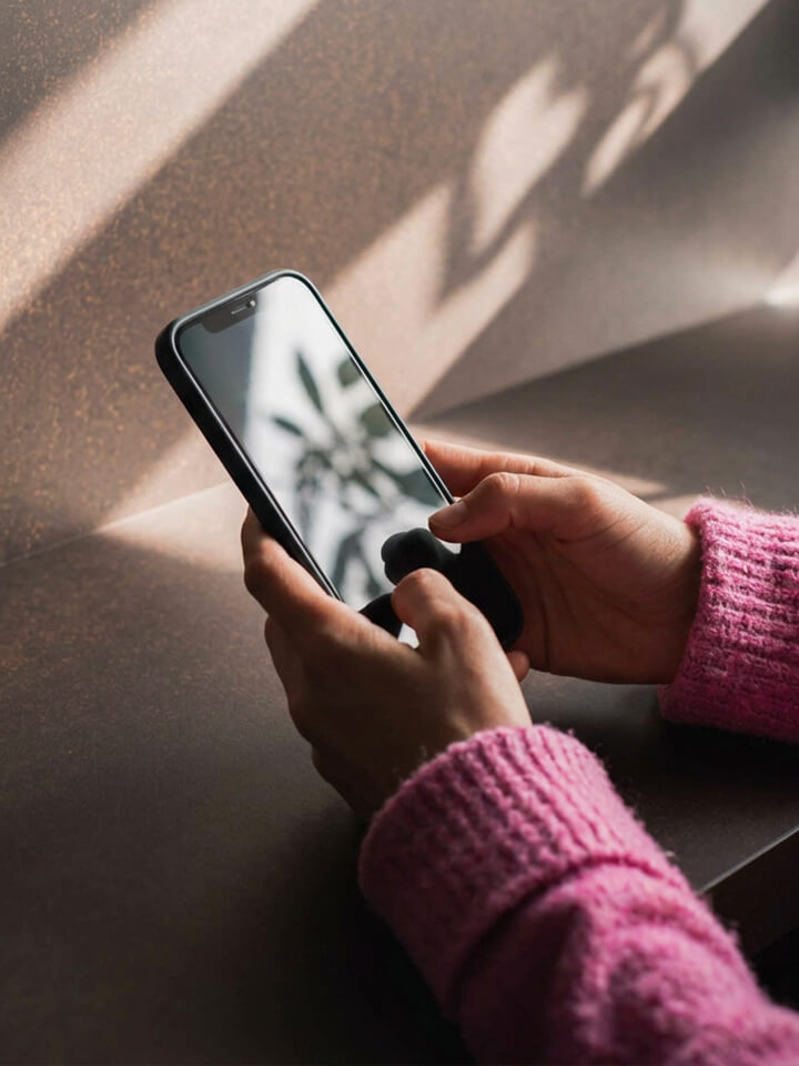 Hände mit rosa Pullover halten Smartphone, Schatten einer Pflanze an der Wand sichtbar.