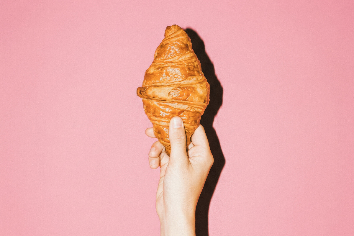 Hand hält frisches, goldbraunes Croissant vor rosa Hintergrund.