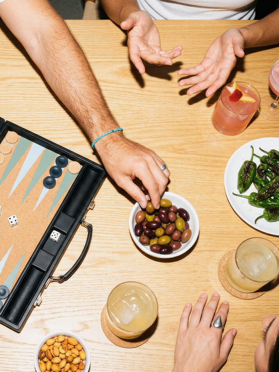 Hände greifen Oliven und Getränke, ein Backgammon-Spiel und Snacks liegen auf einem Holztisch.