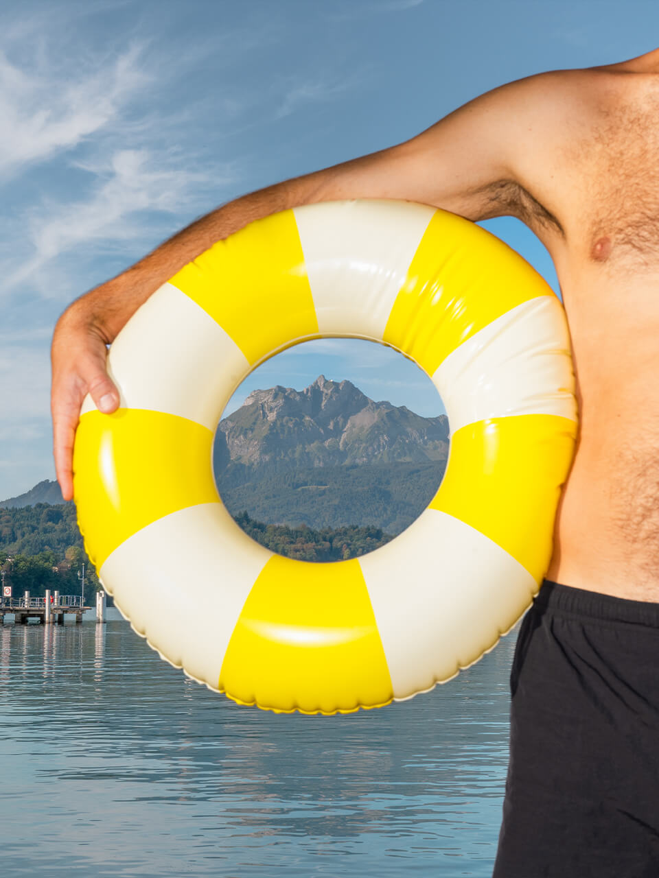 Mann hält gelb-weißen Schwimmreifen vor Berg und See mit blauem Himmel.