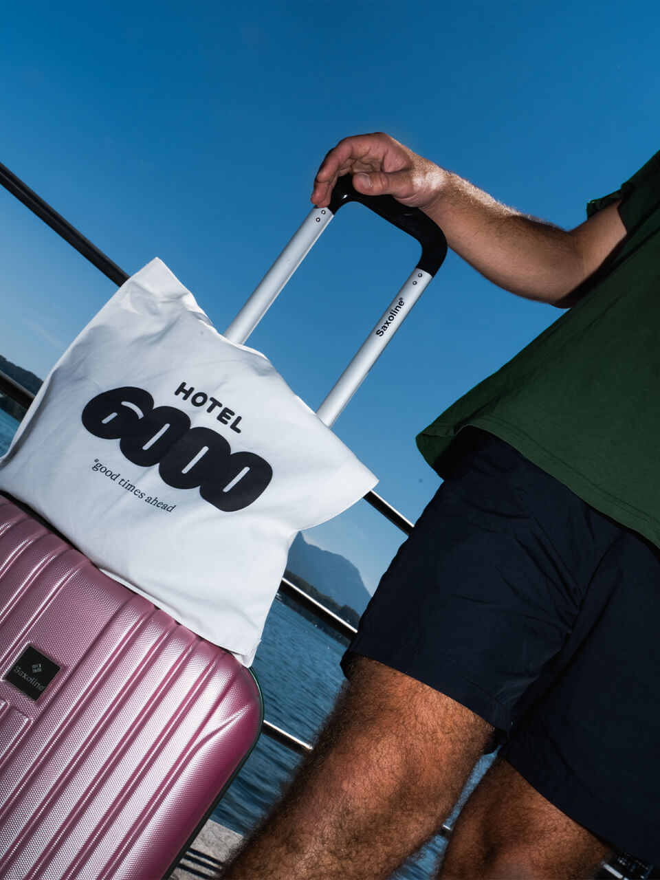 Eine Person hält den Griff eines pinken Koffers mit einer weissen Hotel-6000-Tasche am Wasser unter blauem Himmel.