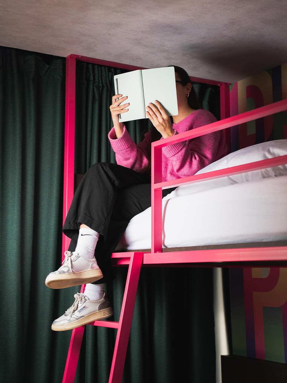 Person sitzt auf einem pinken Hochbett und liest ein Buch, trägt pinken Pullover und Sportschuhe.