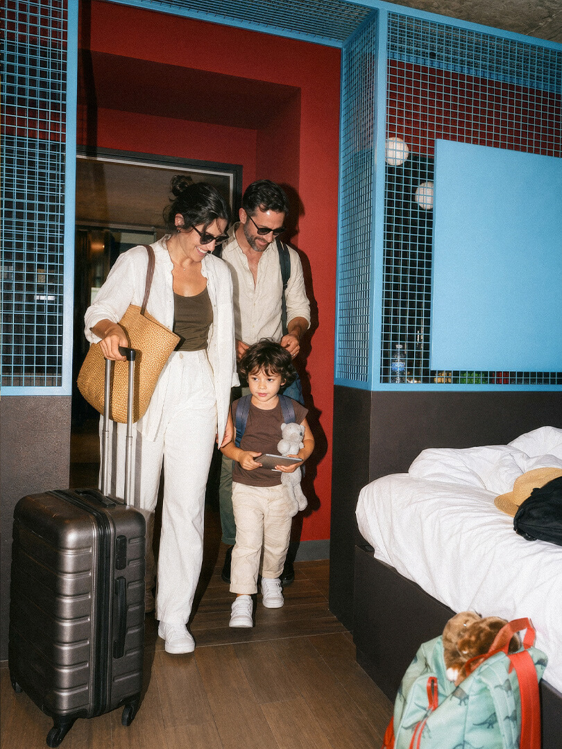 Familie betritt ein Hotelzimmer, die Mutter zieht einen Koffer, das Kind hält ein Stofftier und Tablet.