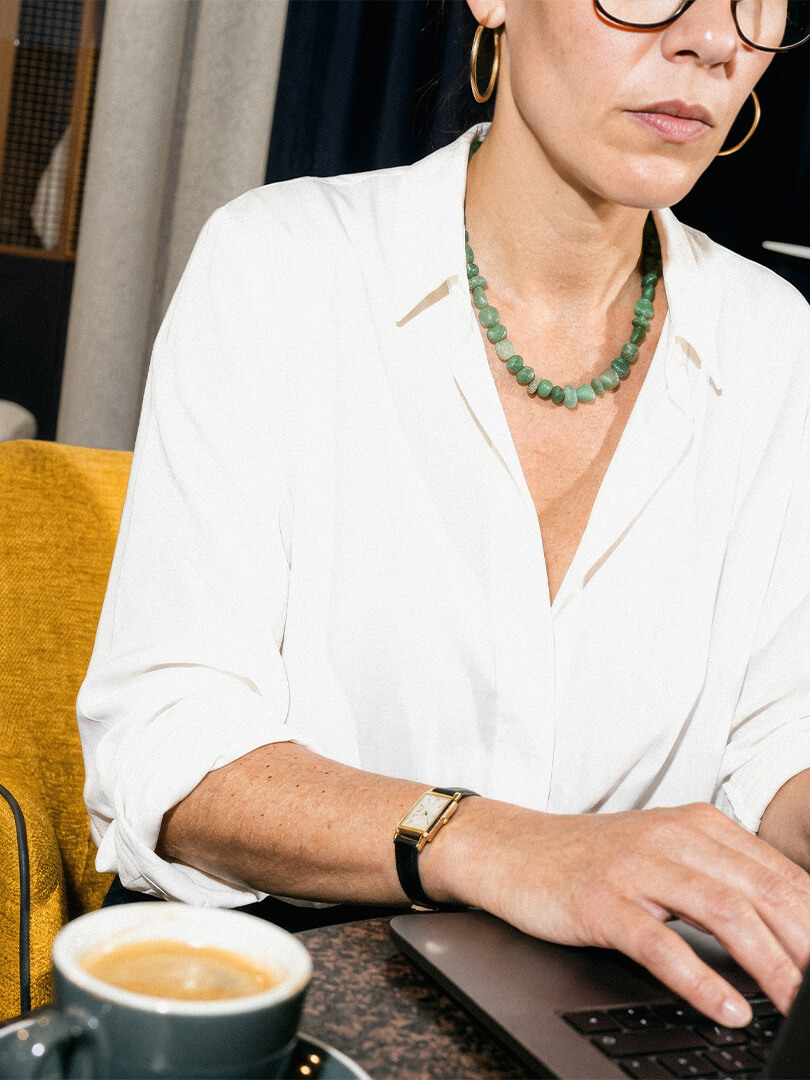 Frau mit weißer Bluse, grüner Halskette und Uhr tippt auf einem Laptop neben einer Kaffeetasse.