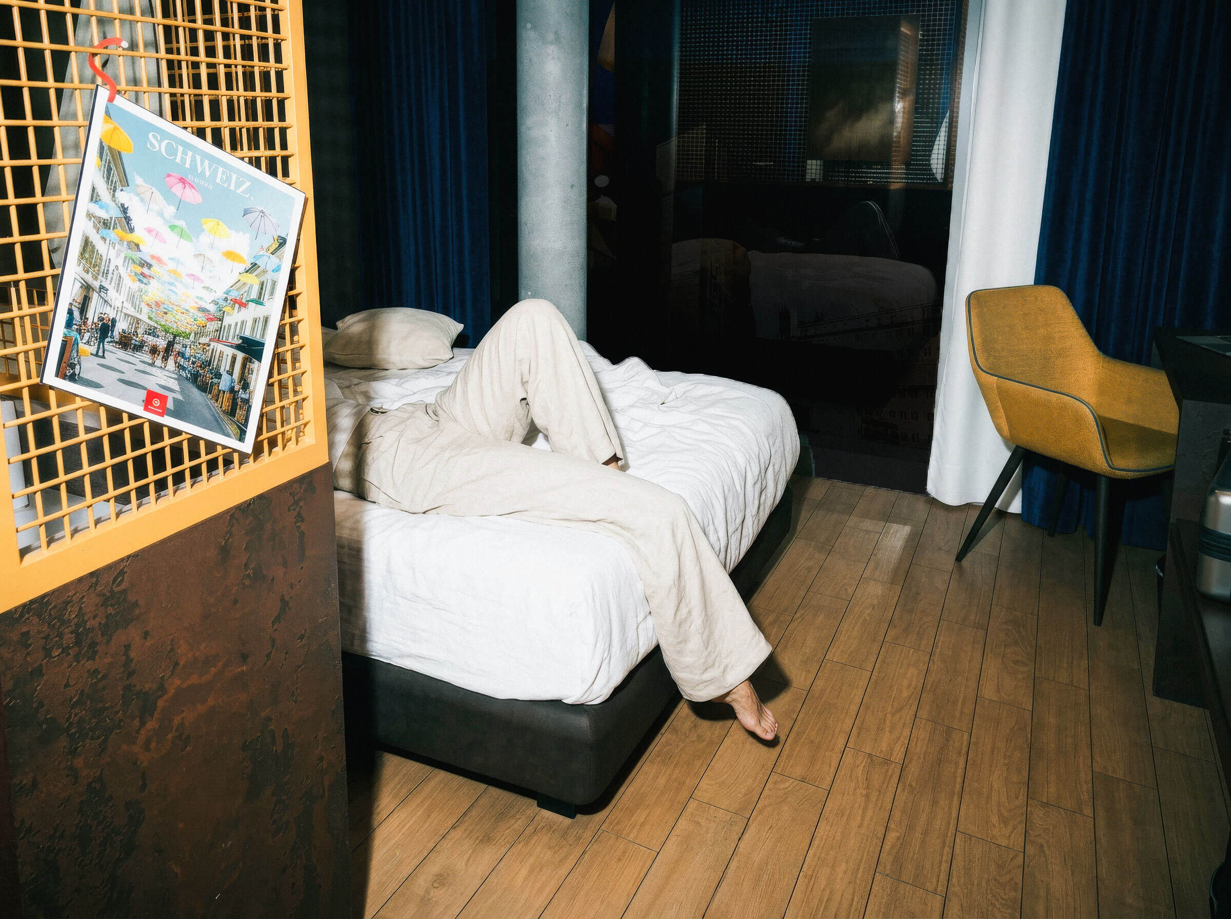 Eine Person in beiger Kleidung liegt auf einem Bett im Hotelzimmer, ein Fuss steht auf dem Holzboden eines modernen Zimmers.