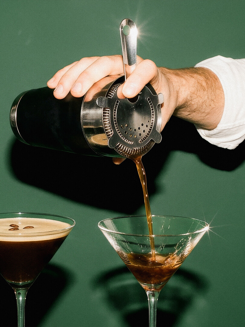 Eine Hand gießt einen Espresso-Martini-Cocktail durch ein Barsieb in ein Martiniglas vor grünem Hintergrund.