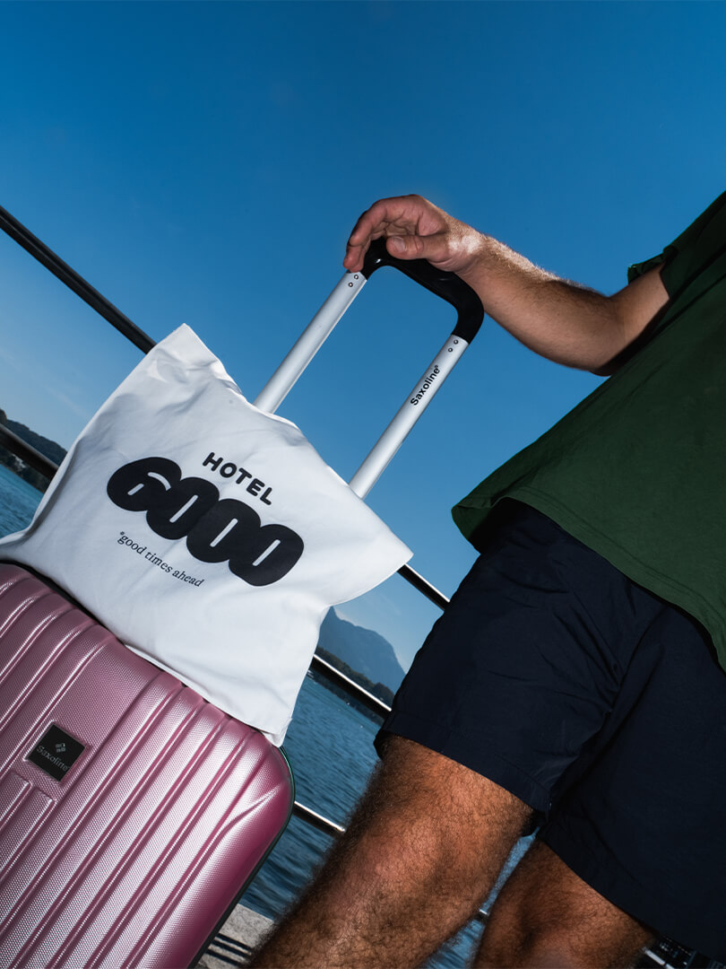 Hand hält Griff von pinkem Koffer mit weißer Tasche Hotel 6000 bei See und blauem Himmel.