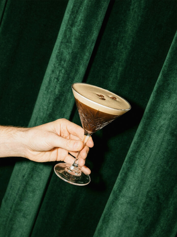 Hand hält ein Cocktailglas mit cremigem Espresso-Martini und zwei Kaffeebohnen vor grünem Vorhang.