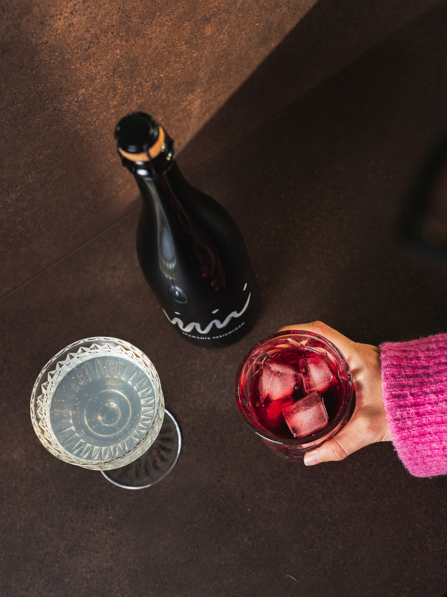 Hand in pink sleeve holding a glass with red drink and ice beside a bottle and a filled cocktail glass.