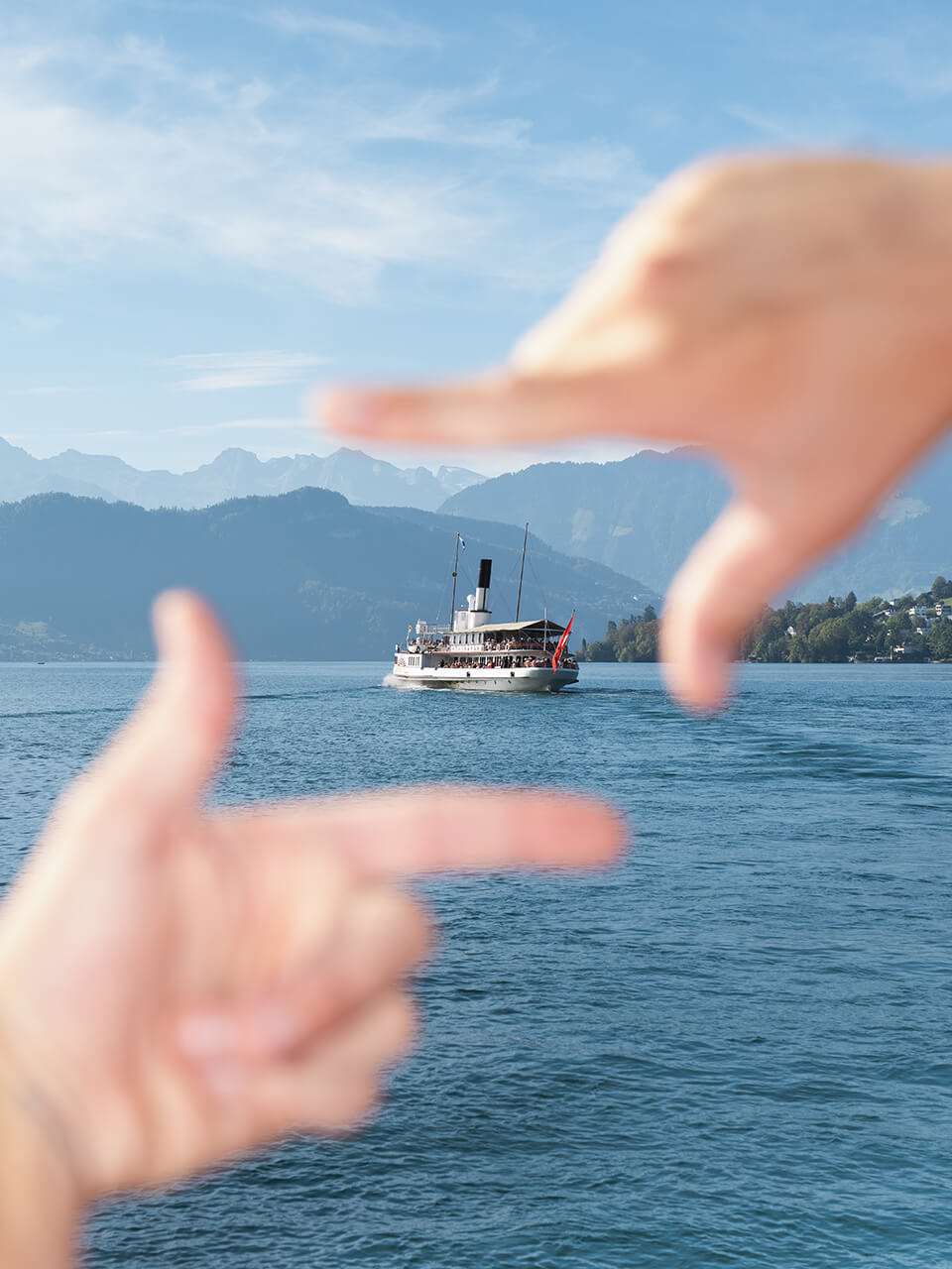 Zwei Hände rahmen ein historisches Dampfschiff auf einem blauen See mit Bergen im Hintergrund.