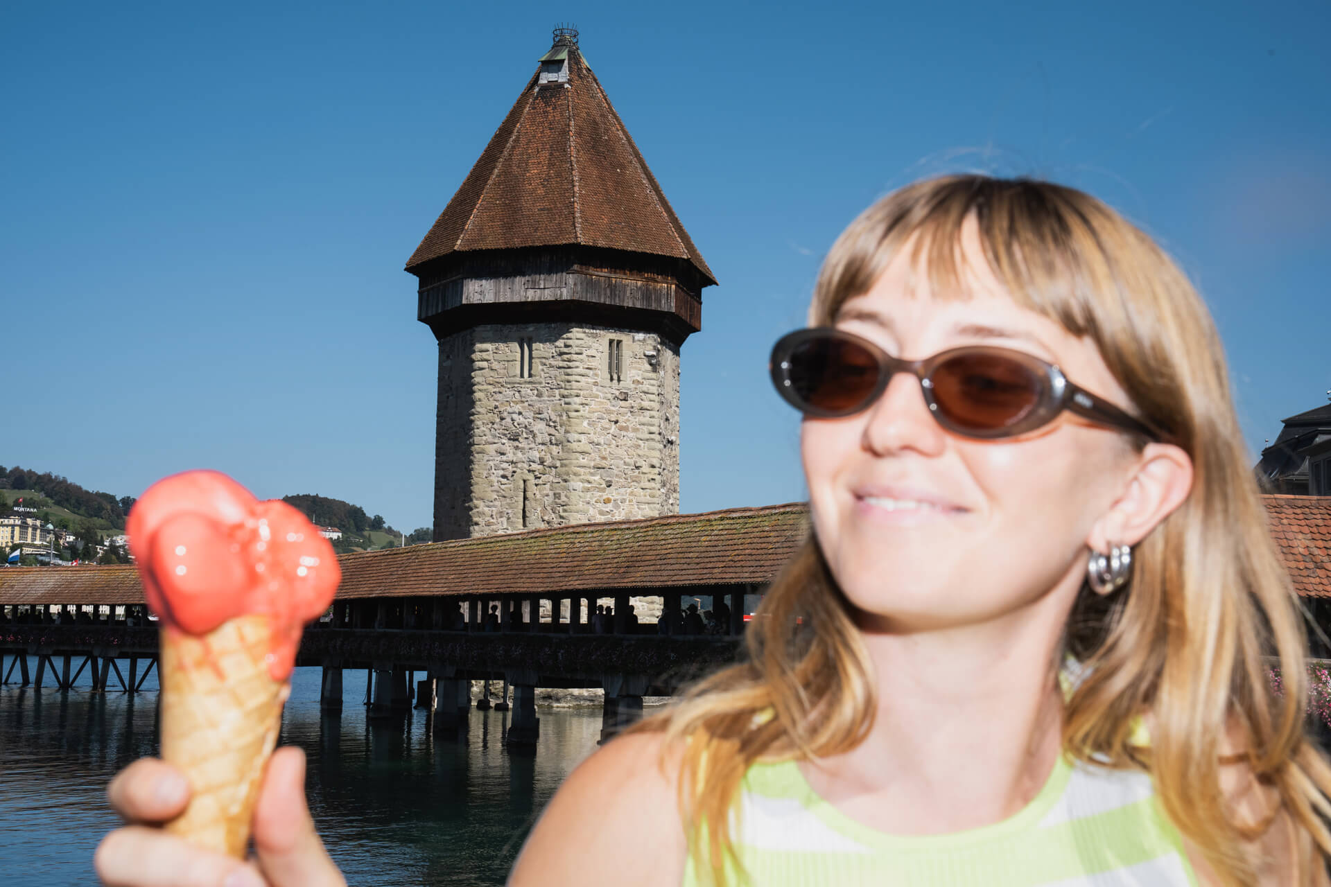 Frau mit Sonnenbrille hält ein rotes Eis, im Hintergrund Lucerne Kapellbrücke und Wasserturm bei Sonnenschein.