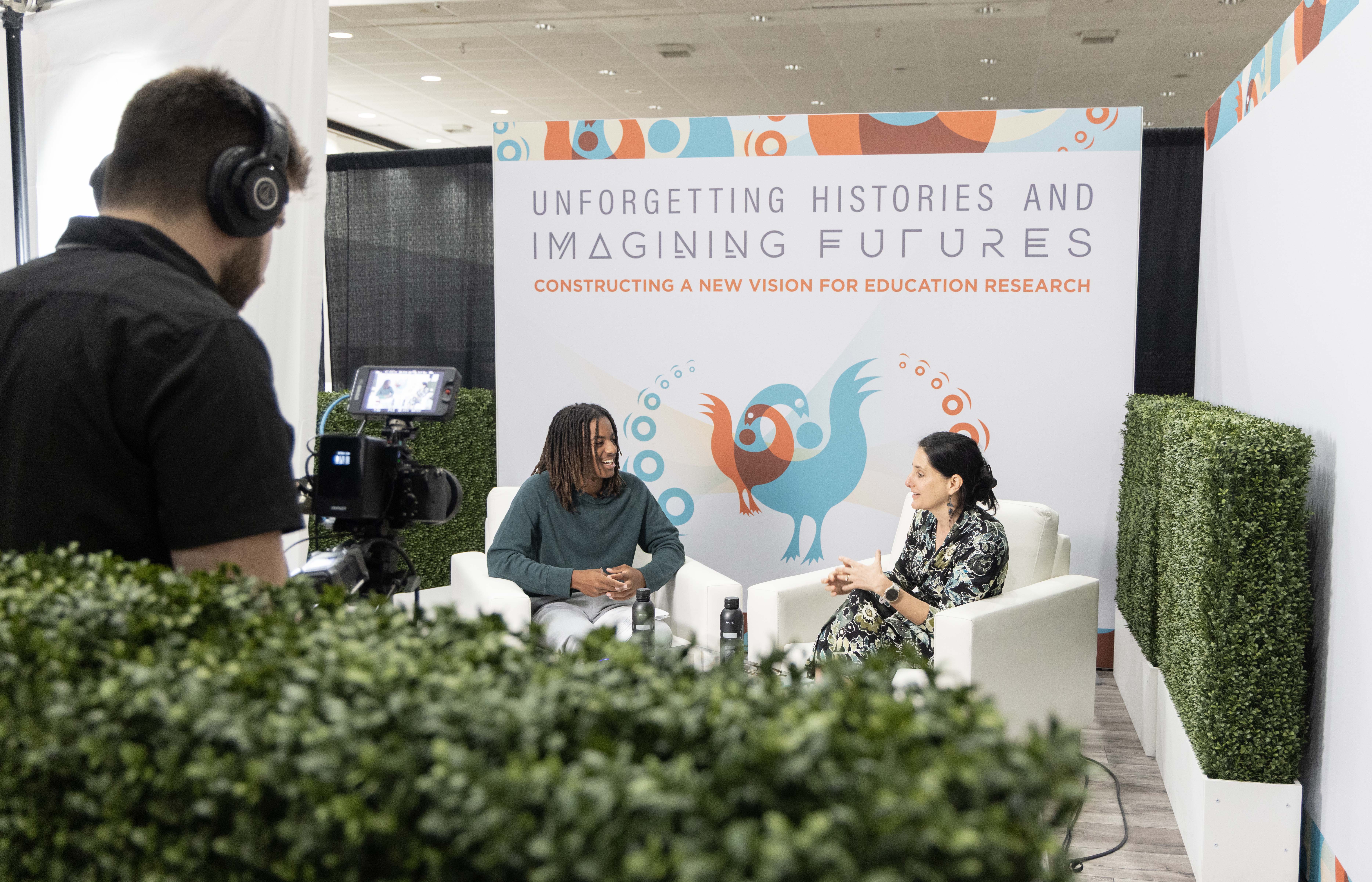 Two people seated for a recorded conversation in front of an AERA conference backdrop